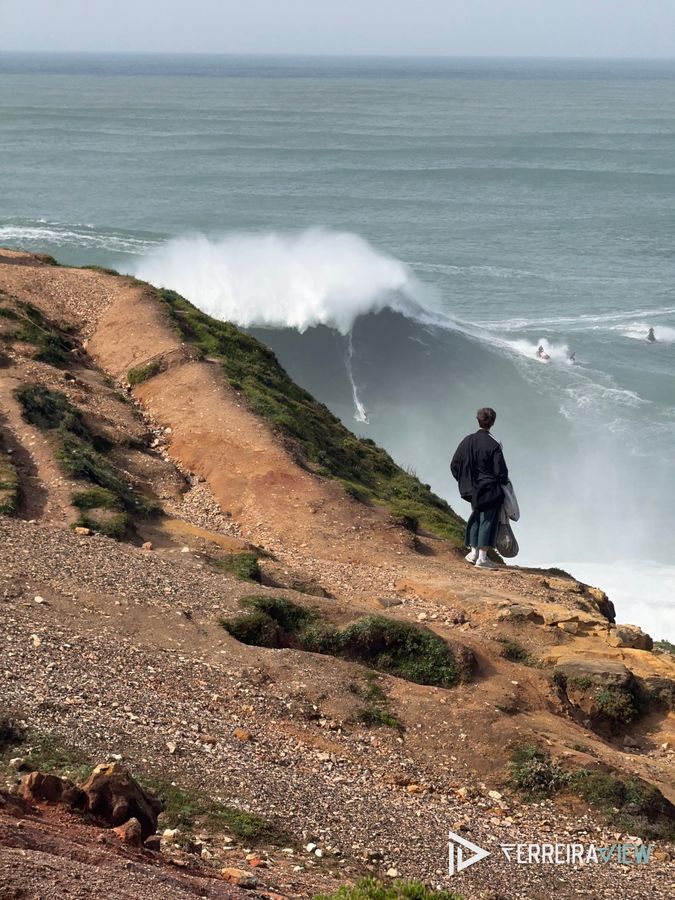 Surfista numa onda gigante na Praia do Norte, Nazaré, com um espectador na falésia